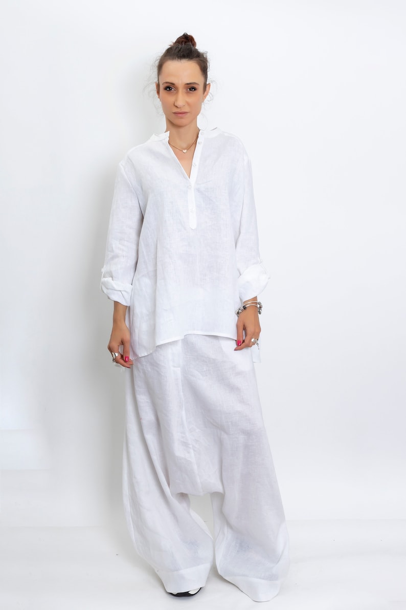 May include: A white linen outfit consisting of a long-sleeved tunic shirt with a mandarin collar and loose-fitting harem pants. The outfit is made of a lightweight, breathable fabric, perfect for warm weather. The model is wearing silver rings and bracelets.