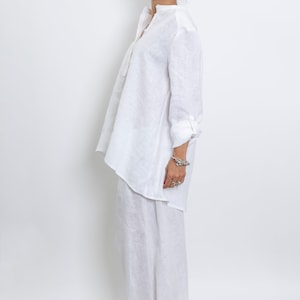 May include: A white linen outfit consisting of a long-sleeved tunic shirt and wide-leg pants. The shirt has a mandarin collar and buttoned cuffs. The person is wearing black-framed glasses and black and white sneakers. The outfit is simple and elegant.