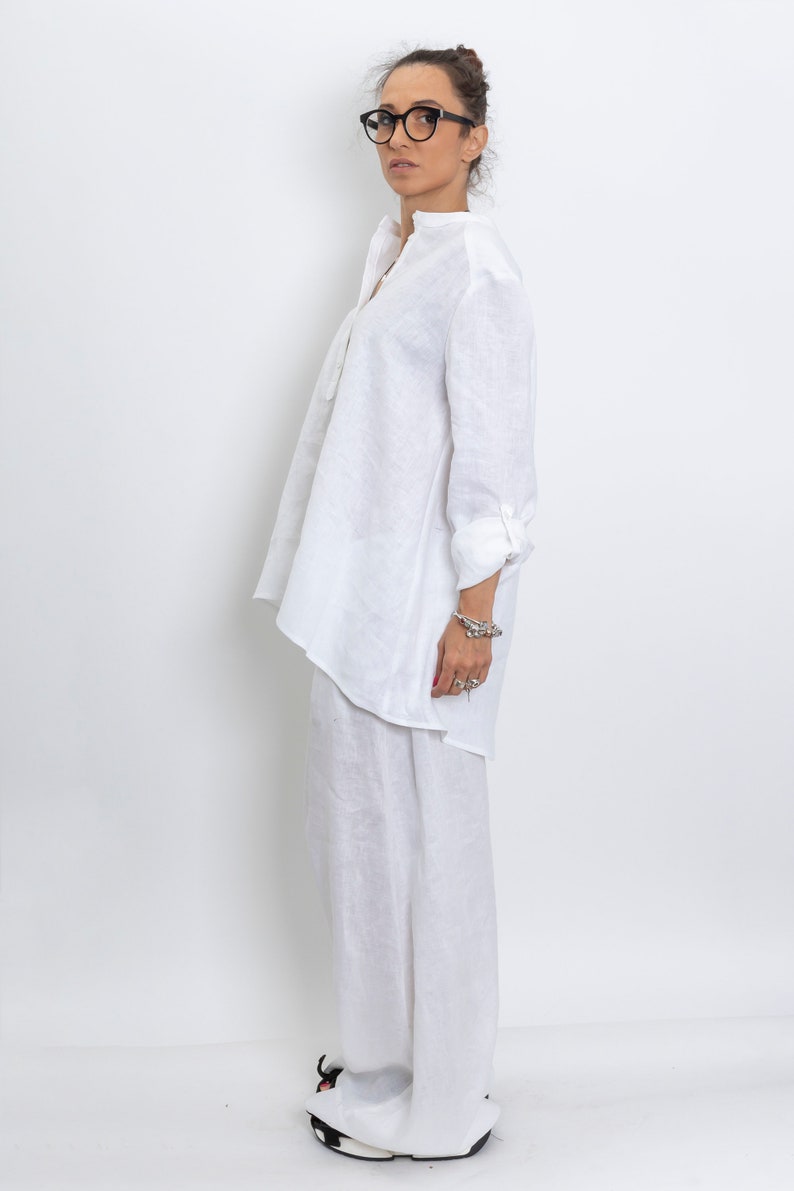 May include: A white linen outfit consisting of a long-sleeved tunic shirt and wide-leg pants. The shirt has a mandarin collar and buttoned cuffs. The person is wearing black-framed glasses and black and white sneakers. The outfit is simple and elegant.