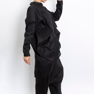 May include: A woman wearing a black, long-sleeved shirt with a ruffled hem, black pants, and black sandals with white soles. She is wearing a black hat and holding it in her right hand.