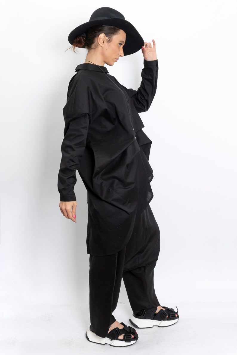May include: A woman wearing a black, long-sleeved shirt with a ruffled hem, black pants, and black sandals with white soles. She is wearing a black hat and holding it in her right hand.