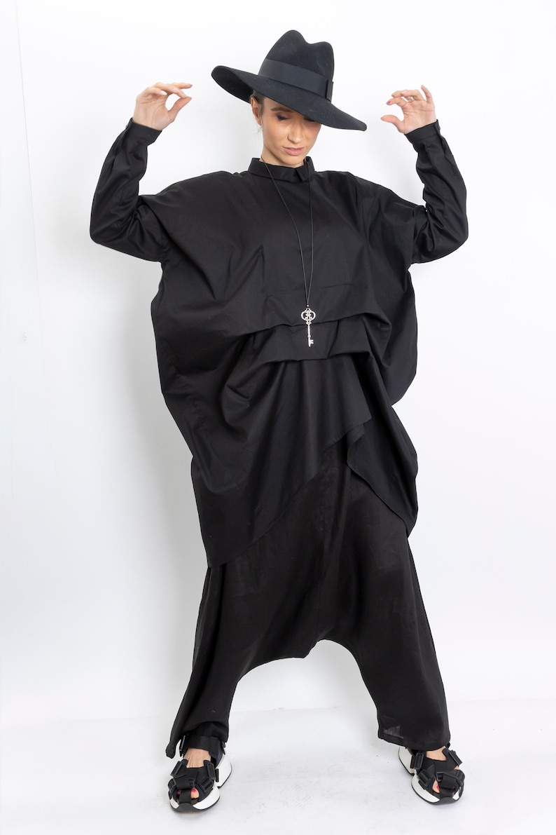 May include: A black wide-leg jumpsuit with a loose-fitting top and a key-shaped pendant necklace. The model is wearing a black fedora hat and black sandals.