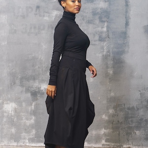 May include: A woman wearing a black turtleneck shirt and a black, flowing skirt. She is wearing burgundy leather boots with laces. The woman is standing in front of a gray textured wall.