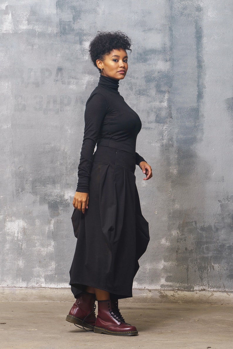 May include: A woman wearing a black turtleneck shirt and a black, flowing skirt. She is wearing burgundy leather boots with laces. The woman is standing in front of a gray textured wall.