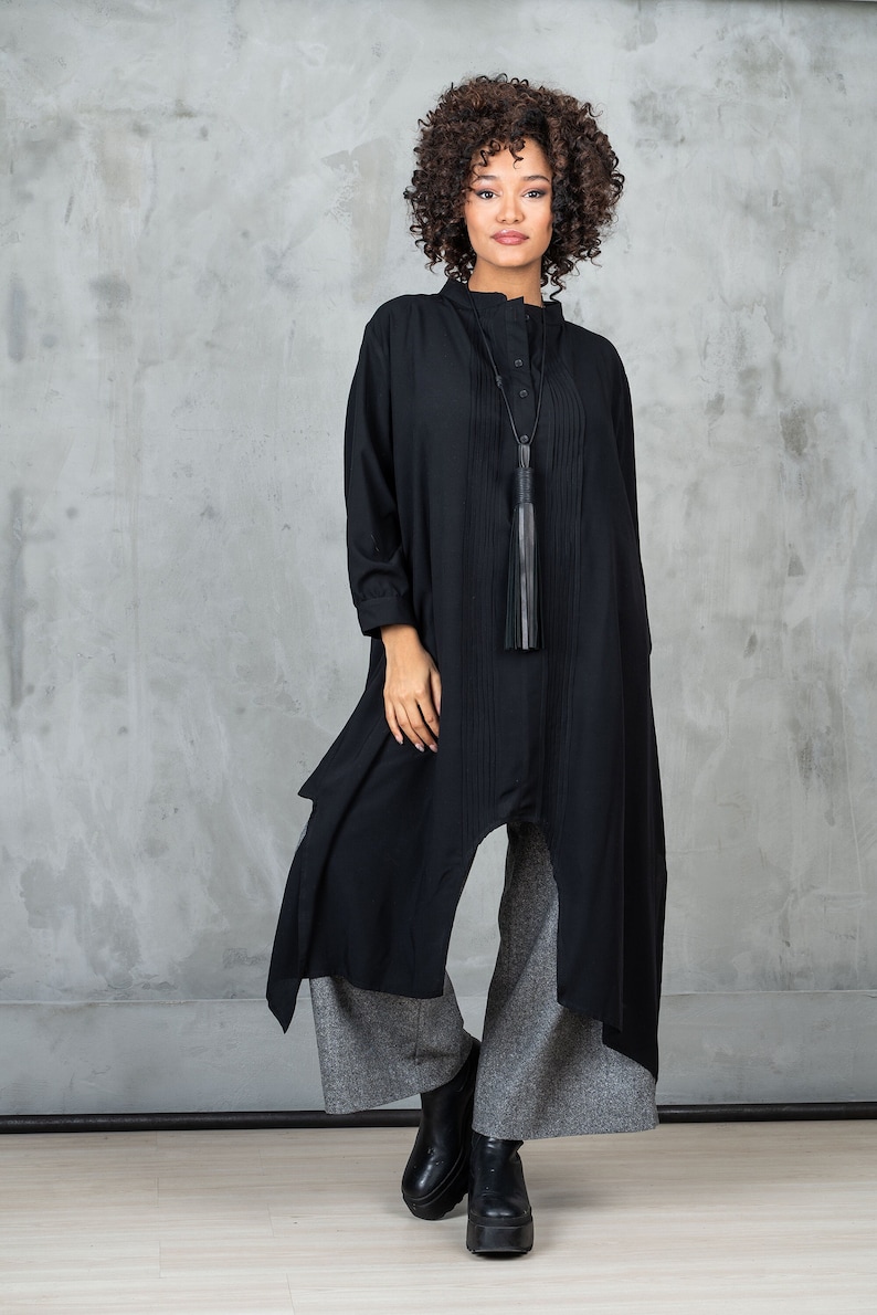 May include: A black long-sleeved tunic with a high neckline and a tassel pendant. The tunic has a pleated front and a flowing, asymmetrical hem. The model is wearing gray pants and black platform shoes.