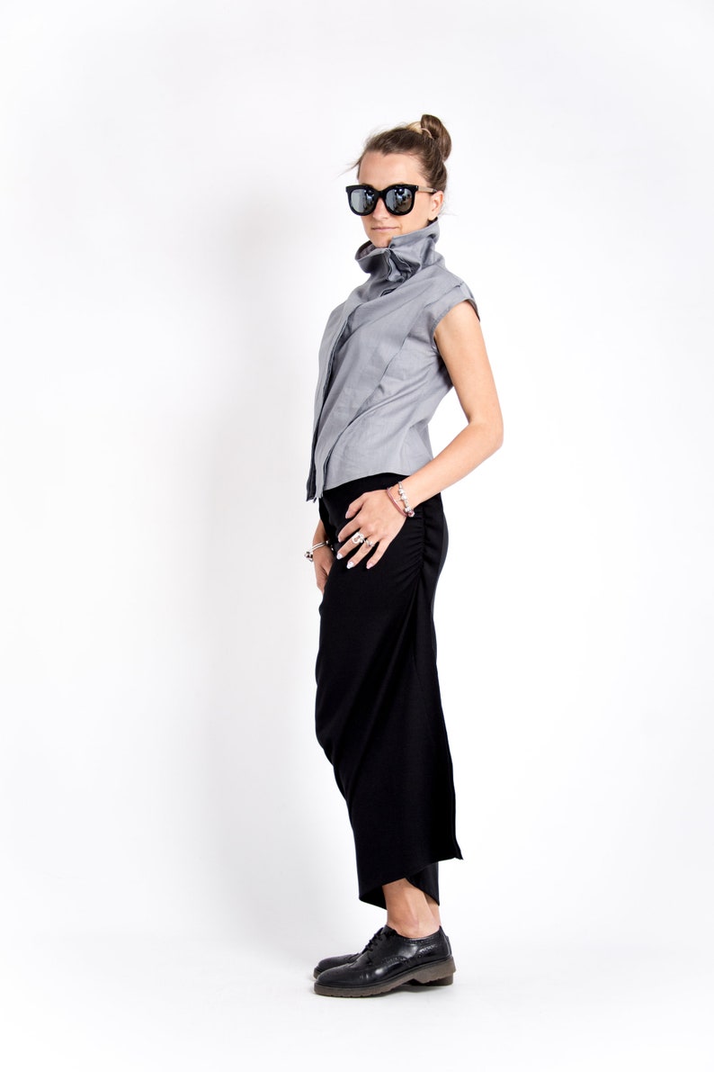 May include: A woman wearing a gray sleeveless top with a high collar and a black skirt. She is wearing black sunglasses and black shoes.