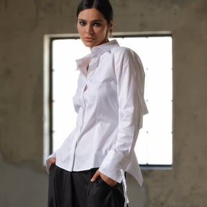 Könnte beinhalten: Ein weißes Langarmhemd mit Knopfleiste und leicht asymmetrischem Saum. Das Hemd ist in eine schwarze Hose mit weitem Bein gesteckt.