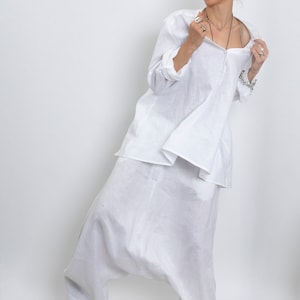 May include: A white linen outfit consisting of a long-sleeved tunic and harem pants. The tunic has a button-down front and the pants have a relaxed fit. The outfit is paired with black sandals.