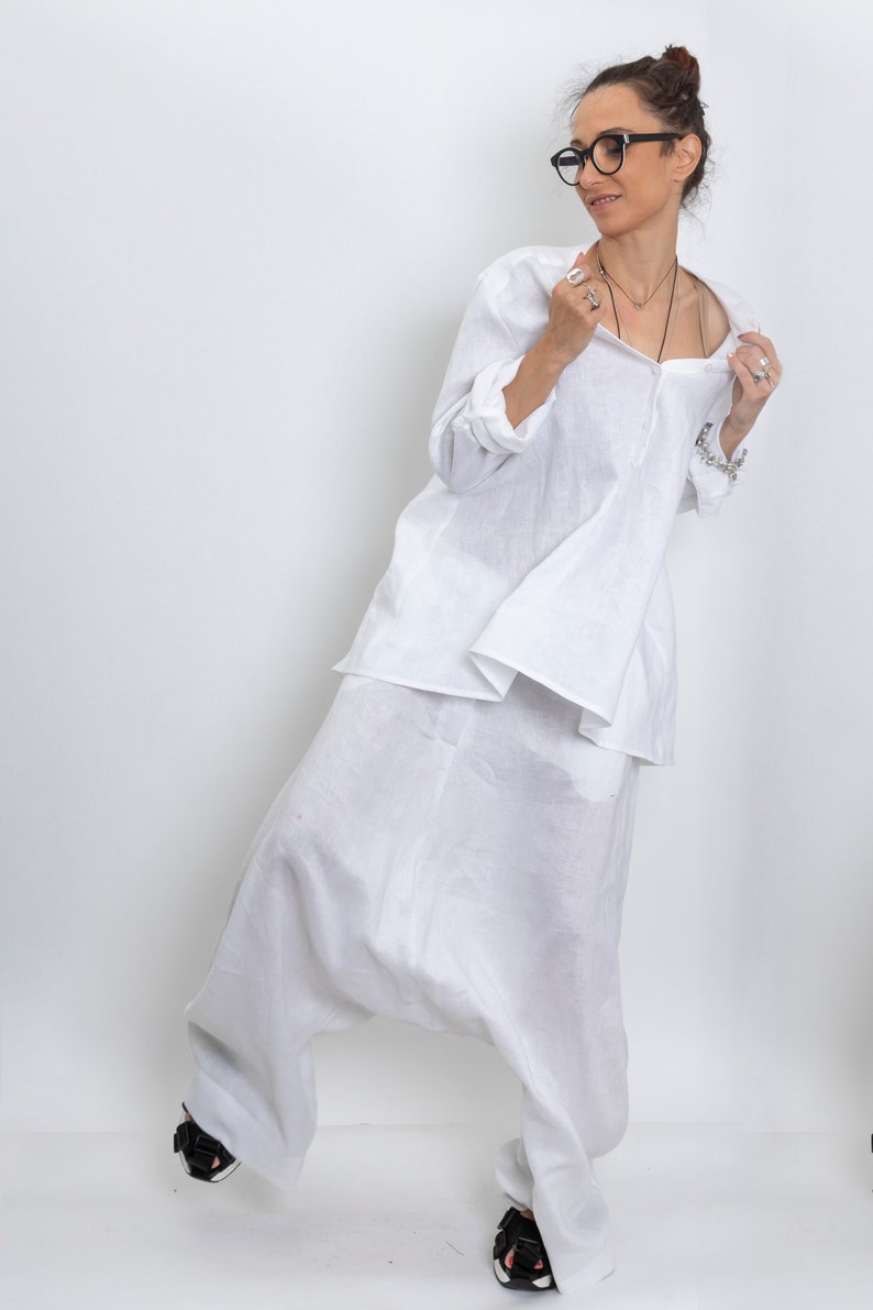 May include: A white linen outfit consisting of a long-sleeved tunic and harem pants. The tunic has a button-down front and the pants have a relaxed fit. The outfit is paired with black sandals.