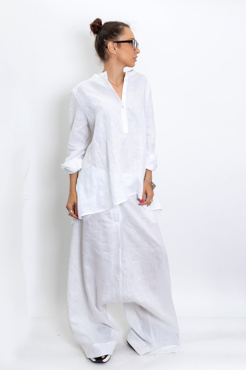 May include: A white linen outfit consisting of a long-sleeved tunic shirt and wide-leg pants. The shirt has a mandarin collar and a button-down front. The pants have a relaxed fit. The outfit is made of a lightweight fabric, perfect for warm weather.