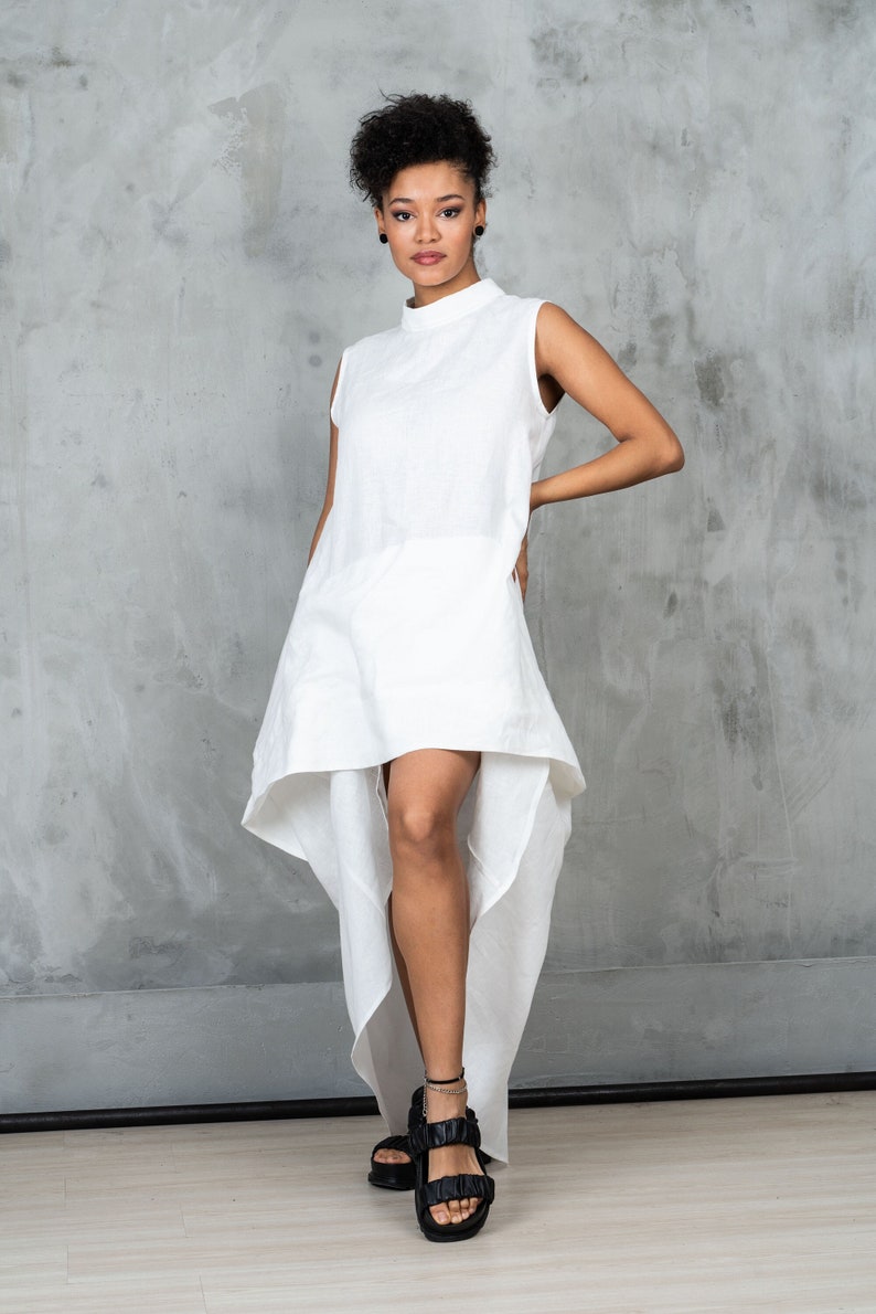 May include: A white linen dress with a high neckline and a high-low hemline. The dress is sleeveless and has a relaxed fit. The model is wearing black sandals.