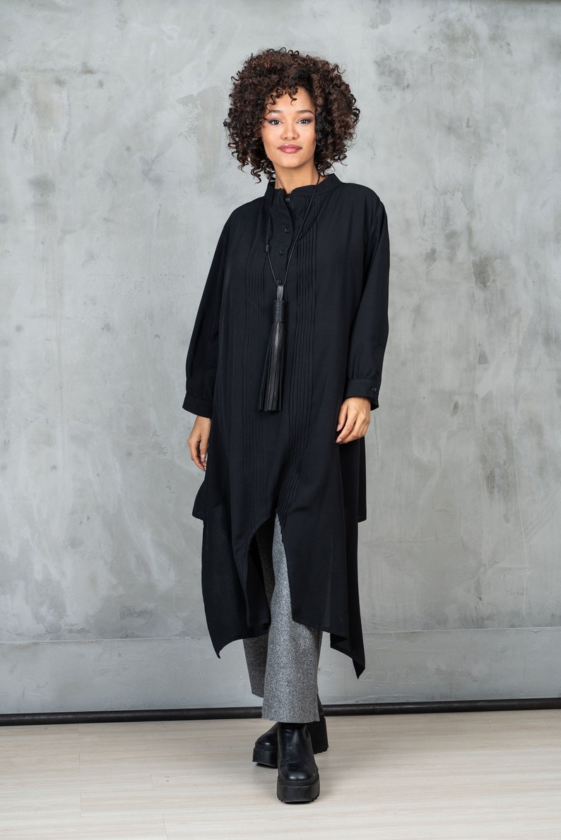 May include: A black long-sleeved shirt with a tassel necklace and a high collar. The shirt has a pleated design and a long, asymmetrical hemline. The model is wearing grey pants and black platform shoes.