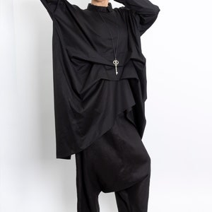 May include: A black, loose-fitting, long-sleeved shirt with a key-shaped pendant necklace. The shirt is layered and has a draped design. The model is wearing black pants and black sandals.