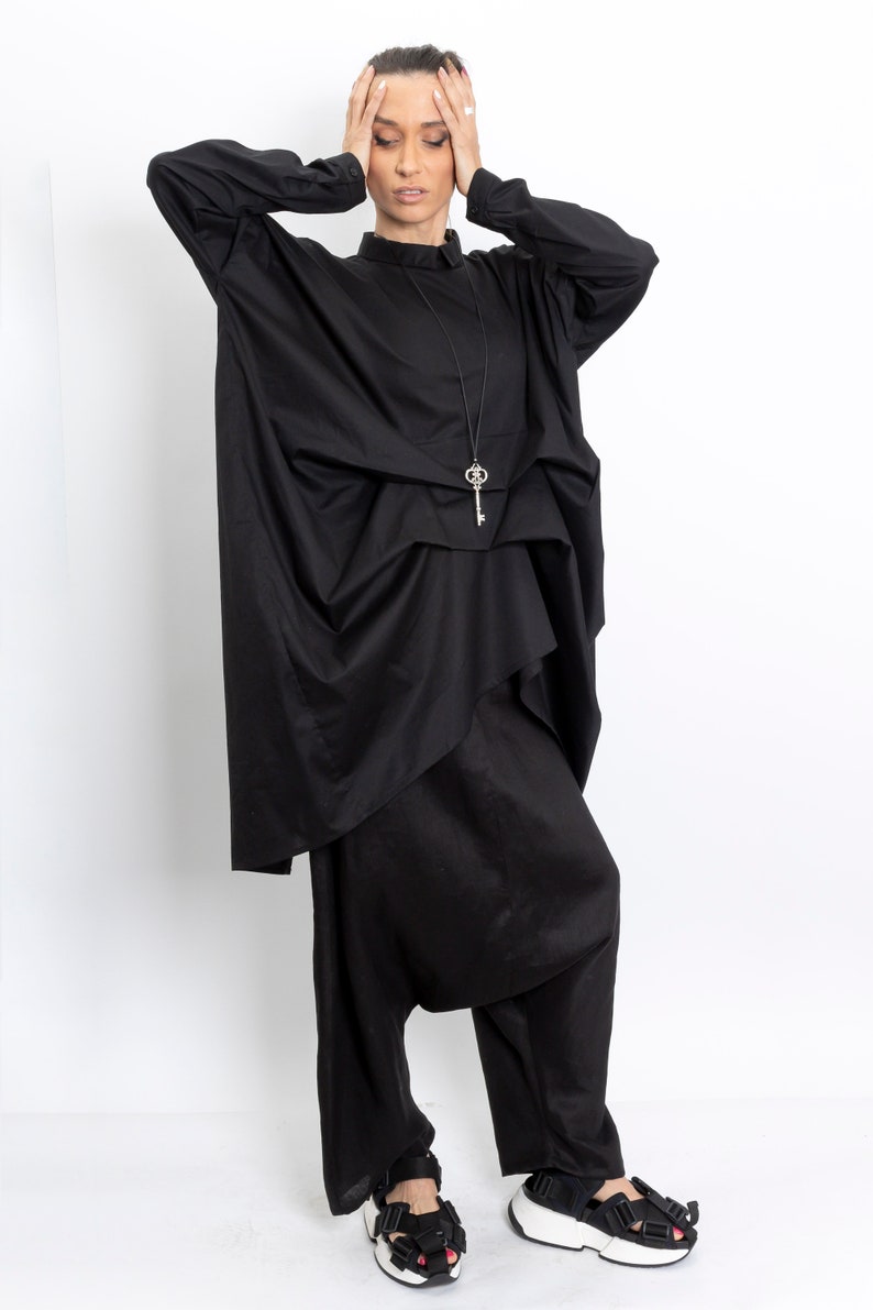 May include: A black, loose-fitting, long-sleeved shirt with a key-shaped pendant necklace. The shirt is layered and has a draped design. The model is wearing black pants and black sandals.