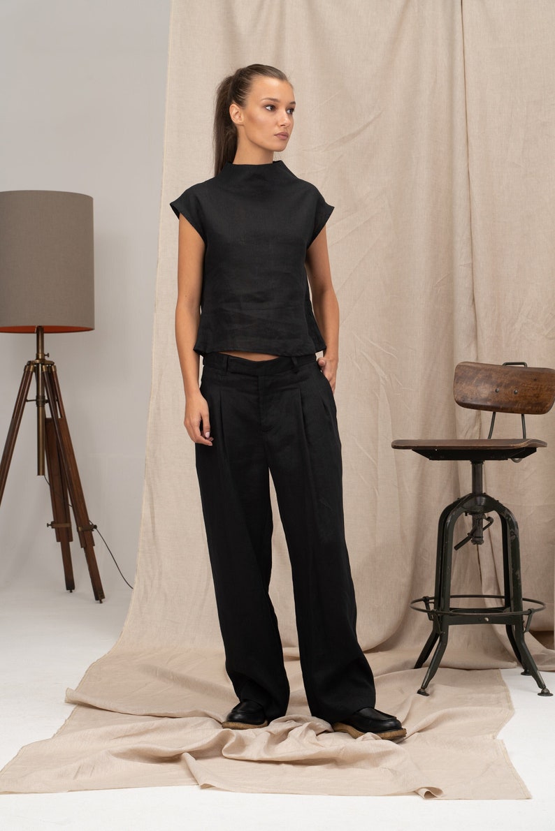 K&ouml;nnte beinhalten: Ein schwarzes Leinenoberteil mit Stehkragen und kurzen &Auml;rmeln, kombiniert mit einer schwarzen Leinenhose. Das Outfit ist im minimalistischen Stil gehalten.