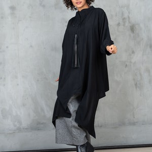 May include: A woman wearing a black, long-sleeved shirt with a tassel necklace and gray pants. She is standing in front of a gray concrete wall.