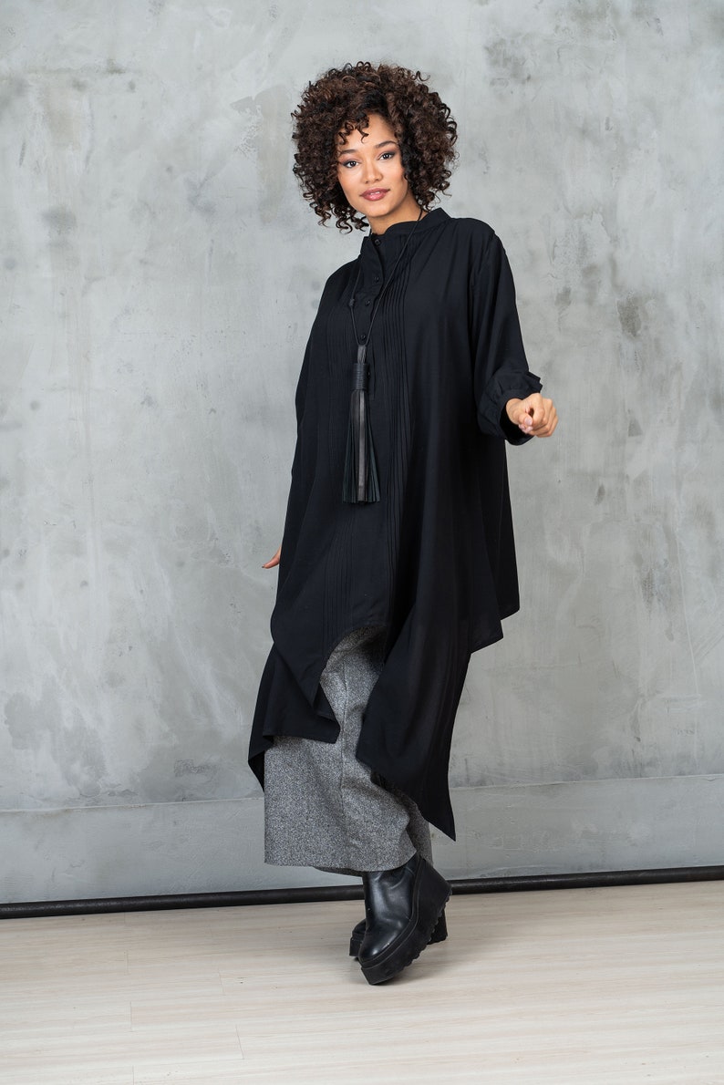 May include: A woman wearing a black, long-sleeved shirt with a tassel necklace and gray pants. She is standing in front of a gray concrete wall.