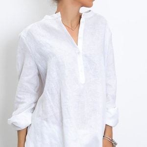 May include: A white linen tunic shirt with a mandarin collar and a partial button-down placket. The long sleeves are rolled up. The shirt has a relaxed fit and is made of a lightweight, breathable fabric.