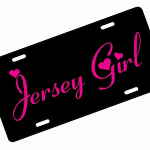 May include: Black license plate with the words "Jersey Girl" in bright pink script, with heart accents. The plate has four mounting holes and a glossy finish. The text is in a cursive font.