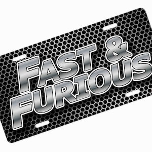 May include: License plate featuring the text "FAST & FURIOUS" in large, silver, 3D-style lettering. The background is a dark grey honeycomb pattern. The plate has four mounting holes.