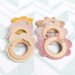 May include: A collection of baby teething rings in various colors. Each ring features a wooden center with a silicone outer ring in shades of pink, beige, and yellow. The names "Penelope" and "Lucy" are engraved on two of the rings.