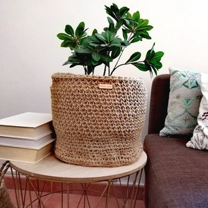 以下が含まれることがあります： 緑の植物が入ったナチュラルなジュート製の植木鉢が、本が積まれた金色の金属製のサイドテーブルの上に置かれています。テーブルは、緑と白の幾何学模様の枕と白い花の模様の枕が2つある茶色のソファの隣にあります。