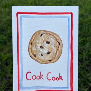 May include: A rectangular card featuring a watercolor illustration of a round chocolate chip cookie centered on a white background. The design is framed by a light blue border and a bold red outer border. The red text "Cook Cook" is printed below.