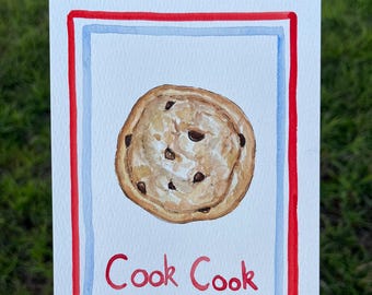 May include: A rectangular card featuring a watercolor illustration of a round chocolate chip cookie centered on a white background. The design is framed by a light blue border and a bold red outer border. The red text "Cook Cook" is printed below.