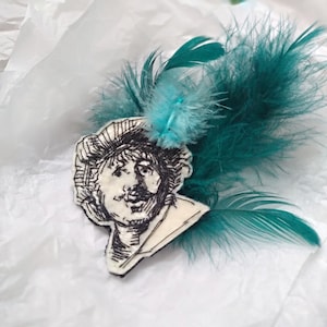 May include: A black and white embroidered portrait of a man with a mustache, set against a white background. The portrait is adorned with a teal feather embellishment.