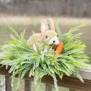 May include: A brown bunny rabbit with white fur around its nose and eyes is holding a carrot in its mouth. The bunny is sitting in a bed of green leafy plants.