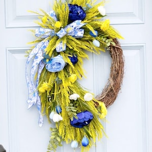May include: A spring wreath with a natural grapevine base, adorned with yellow, blue, and white faux flowers and greenery. A blue and white patterned bow adds a decorative touch. The wreath is displayed on a light blue door.