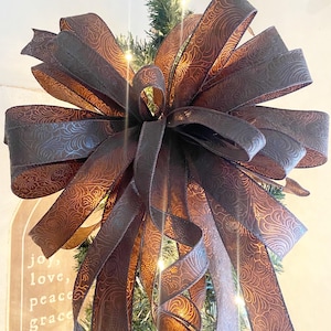 May include: A large brown and gold bow with a floral pattern is tied on top of a Christmas tree.