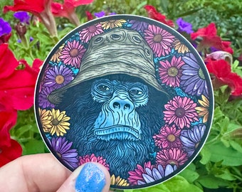 May include: A round sticker featuring a black and blue gorilla wearing a green and brown hat. The gorilla is surrounded by colorful flowers with pink, purple, yellow, and orange petals.
