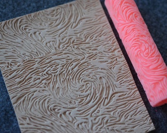 May include: A light brown textured stamp and a pink patterned roller. The stamp has a swirling, organic design. The roller is cylindrical and features a similar swirling pattern. The items are likely used for crafts or art projects.