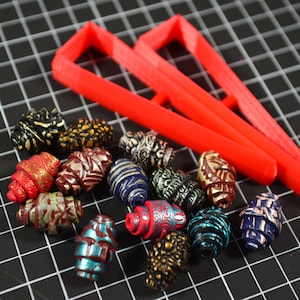 Pode incluir: Uma coleção de contas de argila coloridas e estampadas com uma ferramenta de fabricação de contas de plástico vermelha. As contas são de vários tons de vermelho, azul, verde, dourado e preto.