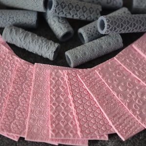 Può includere: Un assortimento di fogli rosa testurizzati e rulli grigi a motivi. I fogli presentano vari disegni in rilievo, tra cui stelle, cuori e spirali. I rulli hanno motivi simili, creando un set per l'artigianato o la decorazione.