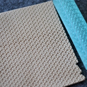 Op de afbeelding: Een lichtblauwe deegroller met een textuurpatroon, gebruikt om een geweven ontwerp op fondant of deeg te creëren. De deegroller ligt naast een stuk fondant of deeg met hetzelfde geweven patroon.