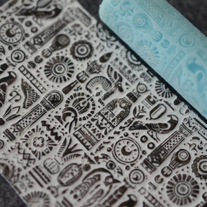 May include: A blue rubber roller with a repeating pattern of geometric shapes and symbols. The roller is used to create a patterned design on a surface.