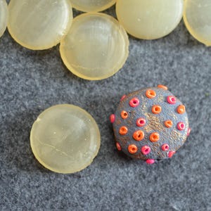 May include: A selection of handmade polymer clay beads. The image shows several translucent, round, light yellow beads and a textured, grey bead with orange and pink accents. The beads are arranged on a grey felt surface.