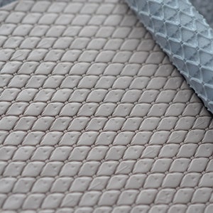 Könnte beinhalten: Nahaufnahme eines beigefarbenen Materials mit Rautenmuster und einem aufgerollten Abschnitt eines ähnlichen, hellblauen Materials. Das Muster besteht aus erhabenen Rauten mit kleinen, kreisförmigen Akzenten an jeder Kreuzung. Die Materialien scheinen weich zu sein.