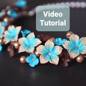 Puede incluir: Primer plano de un collar hecho con flores de arcilla polimérica marrón y beige con centros azules. El collar también presenta hojas marrones y cuentas de imitación de perlas. El texto "Video Tutorial" está en un cuadro azul claro.