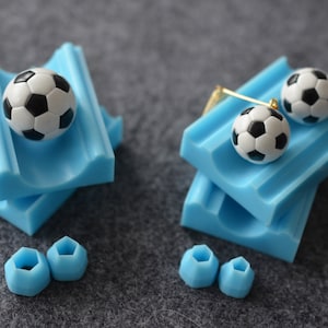 May include: Two blue silicone moulds for making miniature footballs. The moulds are stacked on top of each other. There are three small blue silicone moulds for making other shapes.