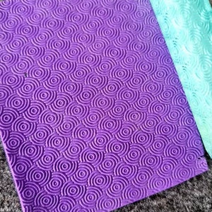 May include: A purple silicone rolling pin with a repeating pattern of concentric circles. The rolling pin is lying on a gray surface.