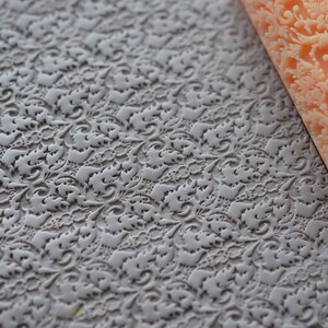 May include: A light gray, textured rolling pin and a matching embossed surface. The rolling pin is a light orange color and features a detailed floral pattern. The surface has a similar floral design, creating a cohesive set for baking or crafting.