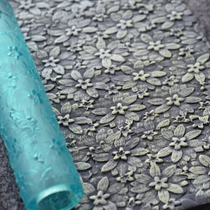 Könnte beinhalten: Eine blaue, aus Kunststoff gefertigte Nudelrolle mit einem Blumenmuster. Die Nudelrolle wird verwendet, um Prägemuster auf Fondant oder Teig zu erstellen.