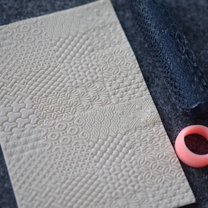 Könnte beinhalten: Eine weiße, strukturierte Rollmatte mit verschiedenen Mustern, ein blauer Rollpin mit strukturiertem Design und ein rosa Silikonring.