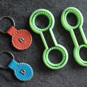 May include: Overhead view of two green bubble wands and two leather keyrings. The keyrings are red and blue, with silver rings and stitched edges. Each keyring has a silver letter. The bubble wands are light green.