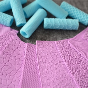 Könnte beinhalten: Ein Set aus 10 Silikon-Nudelholz und 8 Silikon-Matten zum Backen und Dekorieren. Die Nudelhölzer sind hellblau und haben verschiedene geprägte Muster. Die Matten sind lavendelfarben und haben eine Vielzahl von geprägten Mustern.