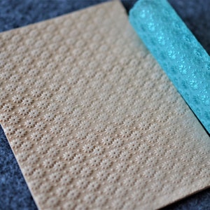 May include: A light blue rolling pin with a decorative pattern of small flowers. The rolling pin is on top of a beige textured surface with a similar pattern.