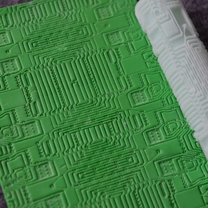 Pode incluir: Um rolo de silicone verde com um design de placa de circuito. O rolo é desenrolado sobre uma superfície cinza.